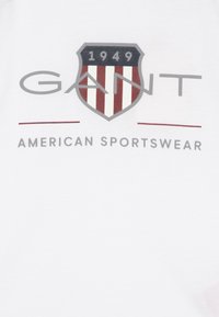 White t-shirt featuring a large shield graphic with red stripes and grey text reading “GANT 1949 AMERICAN SPORTSWEAR” below.