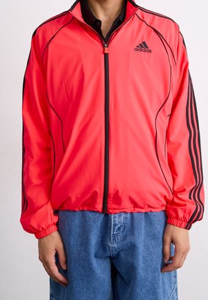 Person trägt eine leuchtend rote Adidas Zip-Up-Jacke mit schwarzen Streifen an den Ärmeln und einem schwarzen Adidas-Logo, kombiniert mit blauen Jeans.