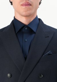 Granatowa marynarka dwurzędowa z klapami wyciętymi, teksturowanym materiałem i czarnymi guzikami. W połączeniu z pasującą granatową koszulą i chusteczką do kieszeni.