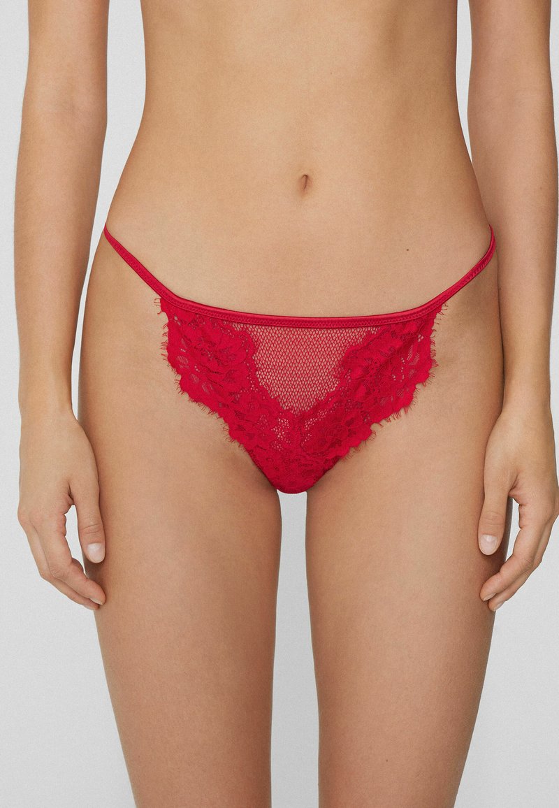 String rouge en dentelle avec design en maille, ornée d'accents floraux et d'une fine ceinture, texture douce et bords délicats.
