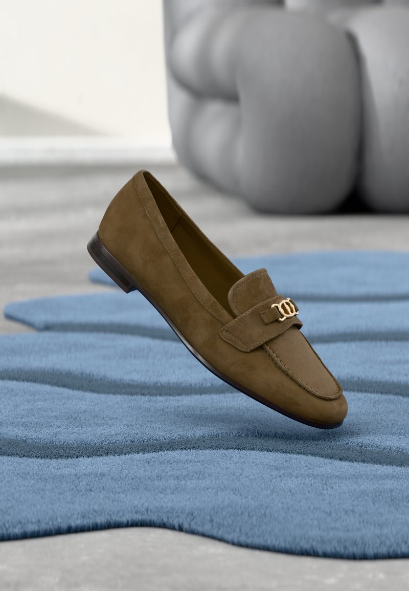 Mocassin en daim marron à texture lisse, doté d'un ornement décoratif en métal doré et d'un bout arrondi, posé sur un tapis bleu.