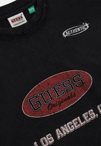 Sweatshirt noir en coton avec un logo ovale rouge "GUESS Originals" usé et le texte "AUTHENTIC", col rond côtelé et étiquette verte.