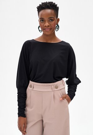 Vrouw met kort krullend haar, gekleed in een zwart shirt met lange mouwen met geplooide mouwen en een beige broek met hoge taille, die lichtjes glimlacht.