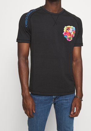 Sort kortærmet t-shirt med farverigt abstrakt ansigtsgrafik på brystet og blå "CARLO COLUCCI" tekst på venstre skulder, båret med blå jeans.