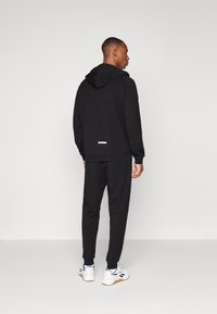 Black hoodie and joggers set, featuring a hood, ribbed cuffs, and a white label detail at the back. Shoes are white with black accents.