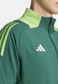Veste de sport verte avec des accents vert clair et trois rayures blanches sur l'épaule. Elle se caractérise par un col haut et une fermeture éclair.