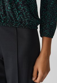 Top nero con pattern astratto verde e vita elastica, abbinato a pantaloni neri aderenti. Primo piano di una mano appoggiata sui pantaloni.