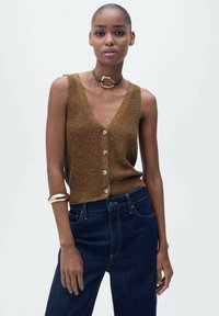 Top sans manches en tricot marron avec un décolleté en V profond et cinq boutons, associé à un jean en denim bleu foncé et un bracelet en or.