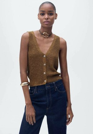 Top senza maniche marrone lavorato a maglia con scollo a V profondo e cinque bottoni, abbinato a jeans in denim blu scuro e un bracciale d'oro.