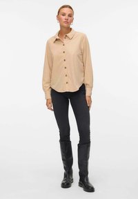 Camicia beige con colletto classico e maniche lunghe, abbinata a jeans neri slim-fit e stivali neri alti fino al ginocchio.