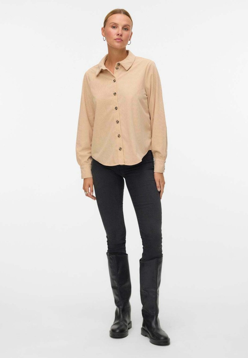 Camicia beige con colletto classico e maniche lunghe, abbinata a jeans neri slim-fit e stivali neri alti fino al ginocchio.