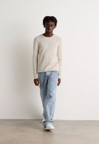 Light beige ribbed sweater, loose light blue jeans, and white sneakers. Model stands against a white background in a neutral space.