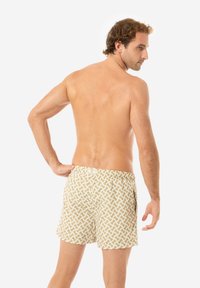 Costumi da bagno di colore beige chiaro con un motivo ondulato bianco, cinturino elastico in vita e una tasca posteriore con chiusura a bottoni.