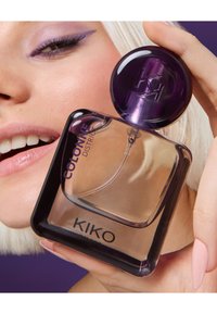 Zbliżenie na rękę trzymającą przezroczystą butelkę perfum KIKO Milano Colonne District w pobliżu uśmiechniętych ust z fioletowym cieniowaniem na powiekach.
