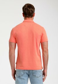 Polo majica s kratkimi rokavi v svetlem koralu z teksturirano tkanino. Ima klasičen ovratnik in ravno rob in je oblečena z svetlo modrimi kavbojkami.