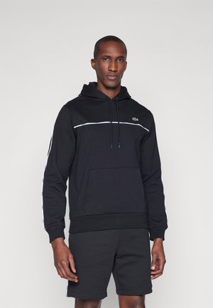 Hoodie preto feito de algodão com um bolso tipo canguru, riscas brancas nas laterais e um pequeno logótipo verde no peito. Combinado com calções pretos.