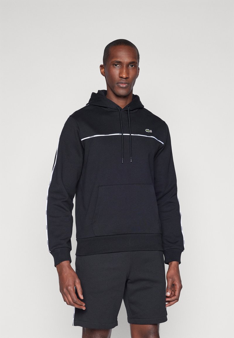 Hoodie preto feito de algodão com um bolso tipo canguru, riscas brancas nas laterais e um pequeno logótipo verde no peito. Combinado com calções pretos.
