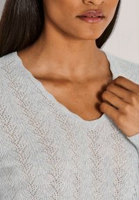 Frau trägt einen hellblauen, strukturierten Strickpullover mit gewelltem V-Ausschnitt und hält einen Stoff nahe am Kragen.