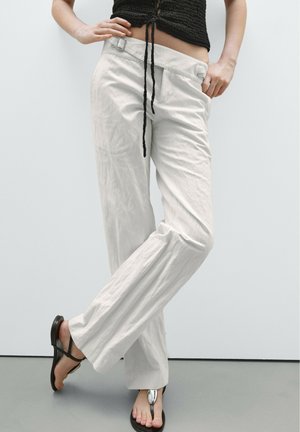 Personne portant un pantalon blanc ample taille haute, un crop top noir et des sandales noires avec un détail argenté, debout les jambes croisées.