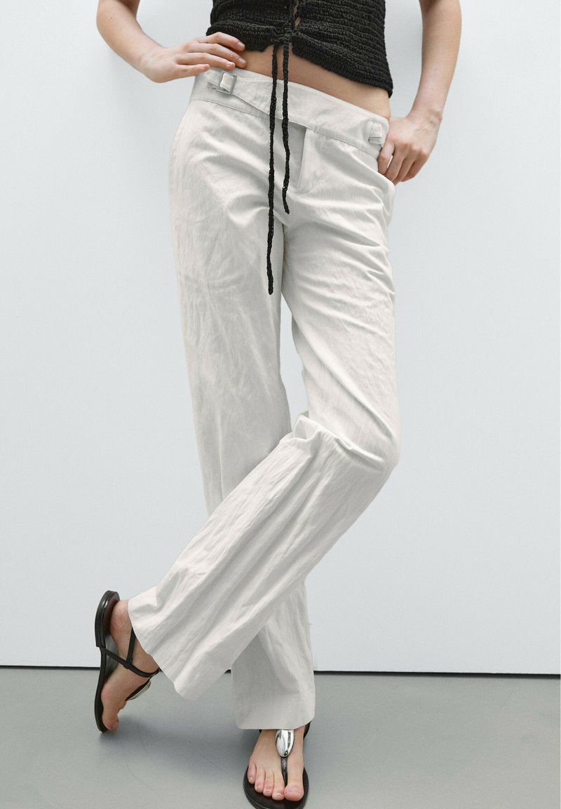 Personne portant un pantalon blanc ample taille haute, un crop top noir et des sandales noires avec un détail argenté, debout les jambes croisées.