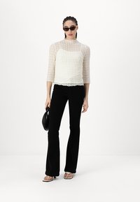 Top alb din dantelă cu mâneci trei sferturi, asortat cu pantaloni negri evazați. Modelul ține o geantă mică neagră, poartă ochelari de soare întunecați și tocuri.