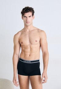 Boxers noirs avec une ceinture grise arborant le logo "LACOSTE", présentant une coupe ajustée et une texture lisse sur un modèle torse nu.