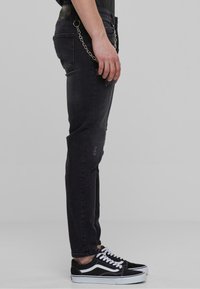 Fekete farmernadrág, slim fit fazonnal, kopott területekkel, fém lánc részletekkel, és fekete-fehér sportcipővel párosítva.