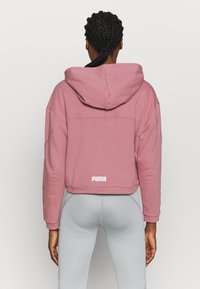 Haut de survêtement rose coupé avec capuche, présentant une couture dans le dos et un logo "PUMA" blanc à la taille. Tissu doux et texturé.
