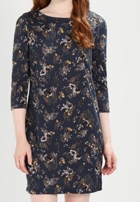 Marinblå klänning med trekvartslånga ärmar, prydd med ett blommönster i nyanser av vitt, gult och brunt. Mjuk, slät spets med rak skärning.