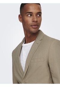 Blazer beige claro en una tela ligera, con cuello con muesca, cierre de un solo botón y bolsillo en el pecho. Llevado sobre una camisa blanca.