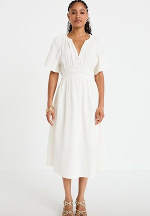 Femme aux cheveux tressés portant une robe midi blanche avec une taille cintrée, des manches flottantes et une fermeture éclair partielle à l’avant, debout devant un fond blanc.