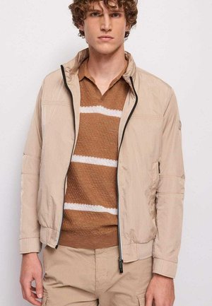 Giovane uomo con capelli ricci che indossa una giacca beige con zip sopra un maglione a righe marrone e bianco, in piedi davanti a uno sfondo bianco semplice.