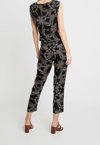 Schwarzer Jumpsuit mit floralem und geometrischem Muster in Weiß- und Goldtönen, mit ärmellosem Oberteil und tapered Beinschnitt. Braune Blockabsatzsandalen.