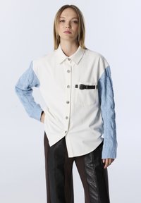 Chemise blanche boutonnée avec manches bleu tricotées en torsades, dotée d'une poche poitrine et d'un détail boucle noire. Le modèle porte un pantalon marron au look cuir.
