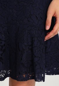 Gonna in pizzo blu scuro con motivo floreale, con una mano che poggia delicatamente sul lato vicino all'orlo.