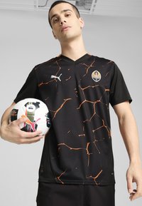 Camiseta de fútbol negra con patrón de grietas naranjas, diseño de cuello en V, que presenta un logo de Puma y el emblema del club. El jugador sostiene un balón con acentos coloridos.