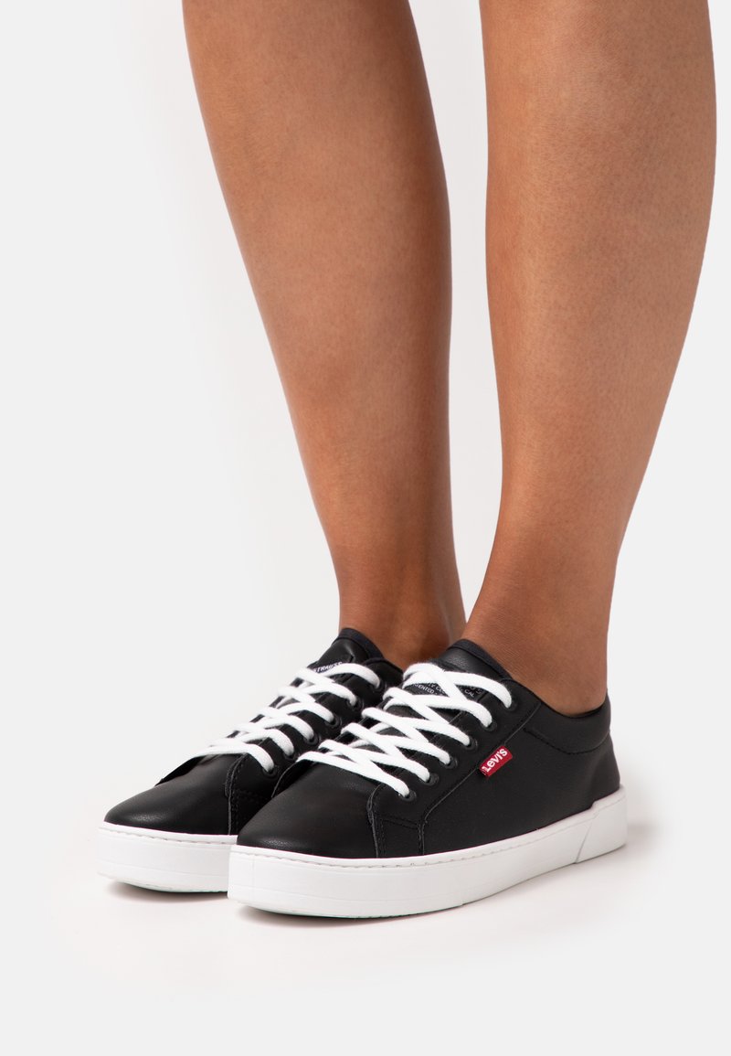 Black leather sneakers with white laces, round toe, and a white rubber sole. Features a small red logo on the side.