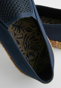 Navyfarvet stof slip-on sko med mesh-overdel, tekstureret espadrille sål og mønstret indersål med bladdesign i mørkeblå på en lys baggrund.
