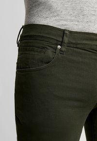 Zbliżenie na ciemnozielone spodnie z przednimi i kieszenią na monety, szlufką na pasek oraz częścią wpuszczonej w spodnie jasnoszarej melanżowej koszuli.