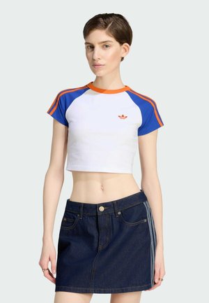 Frau trägt ein weißes Crop-Top mit blauen und orangefarbenen Ärmeln und einen dunklen Jeansminirock mit seitlichen Streifen, steht vor einem einfarbigen Hintergrund.