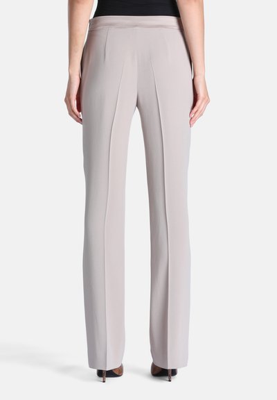 Personne portant un pantalon droit beige clair et des chaussures à talons noires, vue de dos sur un fond uni.