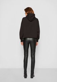 Zwarte hoodie met een grote capuchon, gecombineerd met strakke zwarte leren broeken, die een gladde, glanzende textuur tonen. Model wordt van achteren getoond.