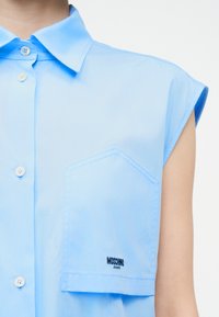 Γαλάζιο αμάνικο πουκάμισο με κουμπιά μπροστά και μεγάλη τσέπη στο στήθος κεντημένη με το λογότυπο "Moschino Jeans".