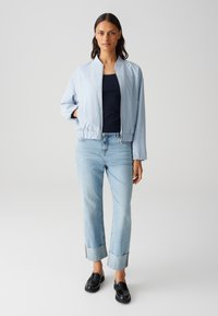 Veste bomber bleu clair avec un ourlet élastique, portée sur un haut bleu marine, associée à un jean bleu clair retroussé et des mocassins noirs.