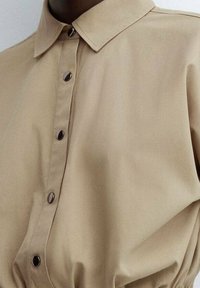 Camicia beige con colletto a punta, con bottoni scuri, tessuto liscio e orlo arricciato per una forma aggiuntiva.