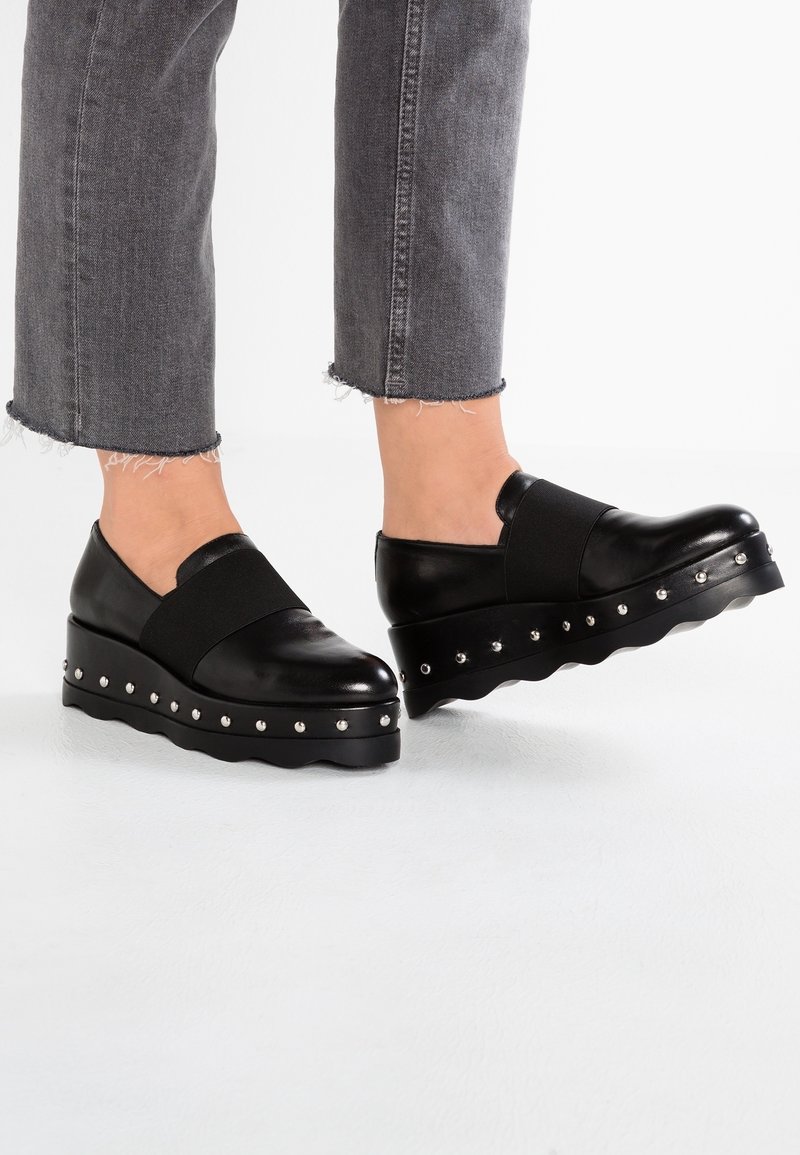 Des chaussures à plateforme en cuir noir présentent une bande élastique sur le dessus, une semelle festonnée et des clous en argent décoratifs le long du bord.