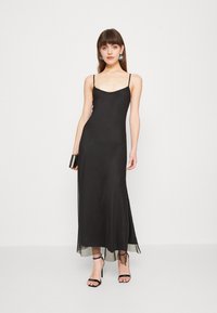 Black sleeveless maxi dress with a fluid silhouette, featuring a smooth material, subtle sheen, and a sheer hemline. Accented with strappy sandals.