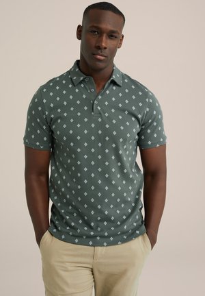 Polo verde con un patrón de diamantes y tapeta con botones. Tejido de algodón suave, mangas cortas. Llevado con pantalones de color beige claro.
