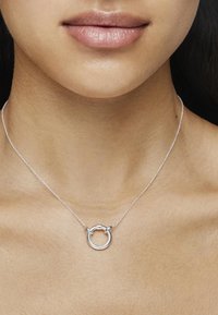Srebrny naszyjnik z okrągłym wisiorkiem, o gładkiej powierzchni i ogniwami, noszony na neutralnym odcieniu skóry. Prosty i elegancki design.