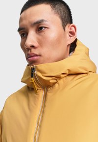 Homme aux cheveux courts portant une veste à capuche jaune moutarde zippée jusqu'au menton sur un fond uni.
